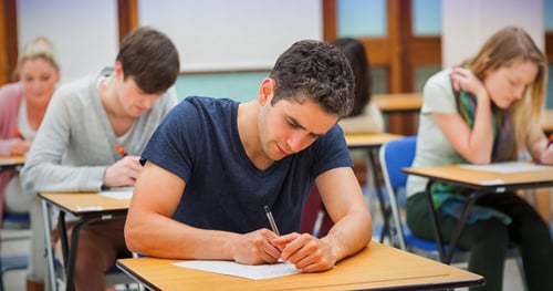 Students seated at wooden desks in a classroom at Highlands College, writing on paper during an exam.