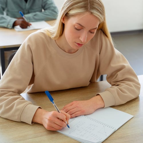 Person writing math equations in a notebook at a desk, with another individual working in the background; equations include algebraic problems and a quadratic expression.
