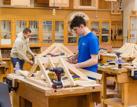 Three individuals working in a woodworking classroom, assembling wooden frames at benches surrounded by tools and materials.