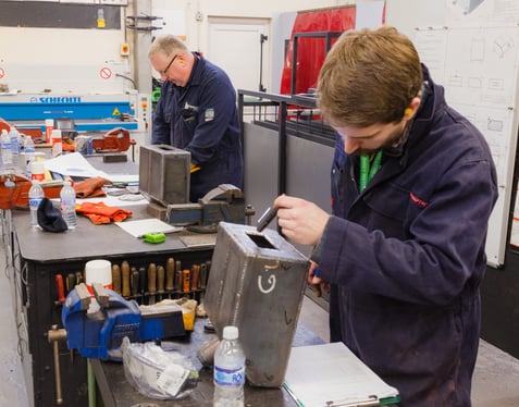 A medium shot of two individuals in protective clothing working in a technical workshop, handling metal boxes on a cluttered workbench with tools, gloves, and diagrams visible in the background.