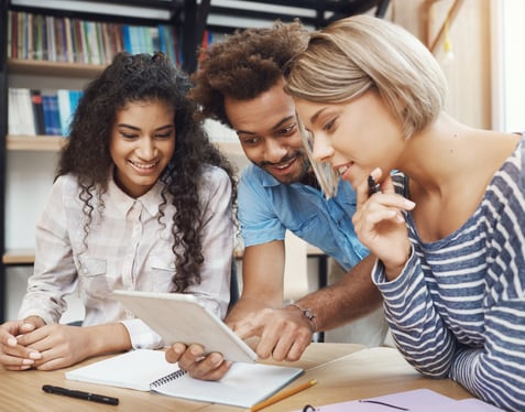 Three people stood around a table, learning on it, looking at a tablet device and smiling.