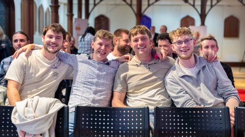 Medium shot of four individuals seated in chairs within a grand hall smiling at the camera with their arms around each other's shoulders.