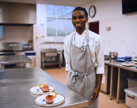 Smiling chef in white coat and apron standing in commercial kitchen with plated desserts