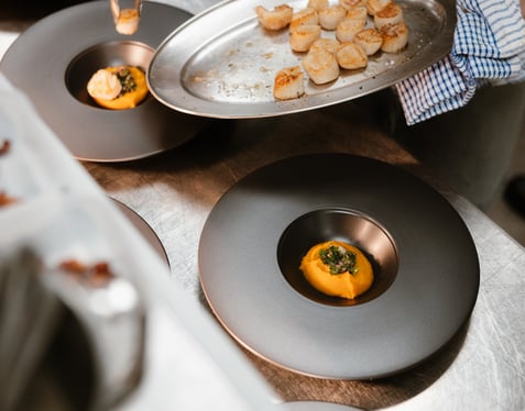 Culinary student at Highlands College plating seared scallops with orange puree and green garnish on black plates in a professional kitchen.