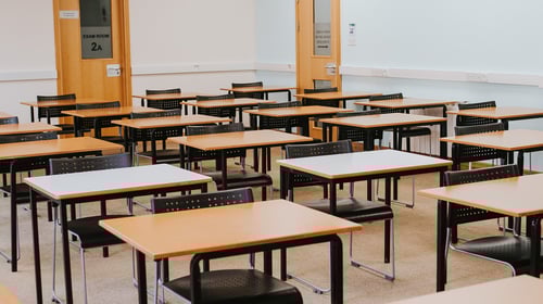 Empty classroom with rows of desks and black chairs arranged in a grid, and a door labelled 'Exam Room 2a' visible in the background.