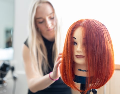 Hairdressing student at Highlands College styling a red wig on a hair-tools mannequin head during salon training.