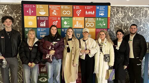 A medium shot of eight individuals looking at the camera standing in front of a small board of coloured squares with several languages written on each square.