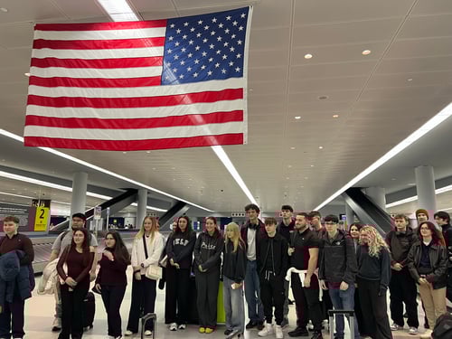 UCJ students in an airport in New York standing underneath an American flag.