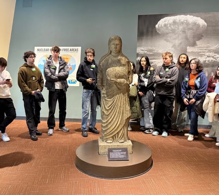 UCJ students in a room standing around a stone statue of a woman holding a lamb. On the walls behind them are photos of the first atomic bomb tests.