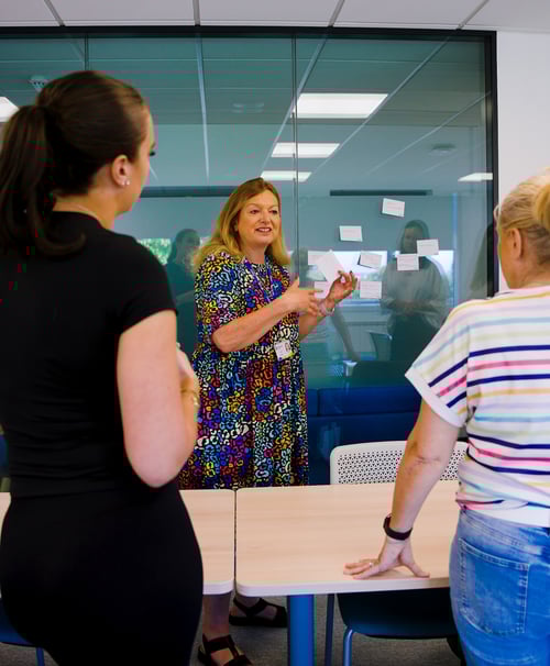 Teacher talking to two adult female students in the classroom, teacher is facing the camera and students are looking at her.