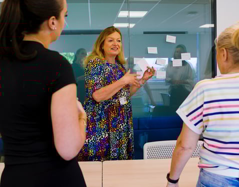 Teacher talking to two adult female students in the classroom, teacher is facing the camera and students are looking at her.