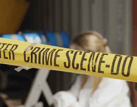 A medium close-up of yellow "Crime scene - do not cross" tape. A blurred individual is crouched down behind the tape looking off to the left.