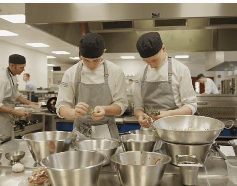 A medium shot of a kitchen filled with chefs. Two of the chefs stand in front of a metal table covered in mixing bowls and cooking ingredients.