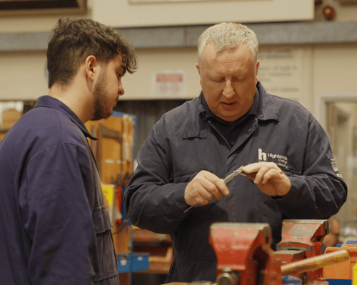 A medium shot of two individuals in a workshop environment. One of the individuals is showing the other two small metal tools.