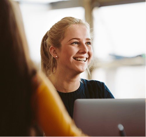 A young woman with blonde hair smiles while looking at someone across a laptop in a bright indoor setting.