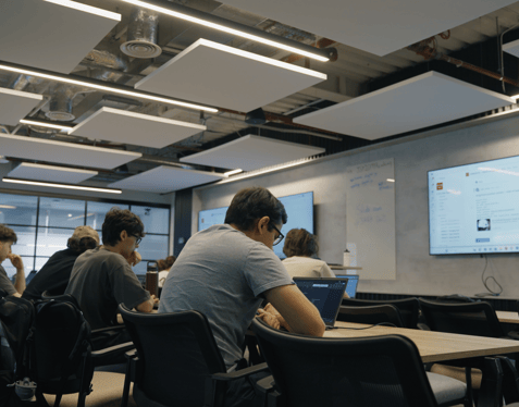 A medium wide shot of students sitting at desks in front of TV screens. There are laptops on the desks with notes on their screens.