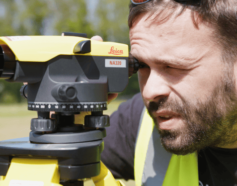 A medium close up of an individual wearing a high-visibility jacket looking through a construction measuring tool.