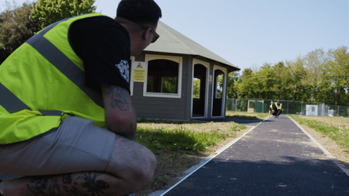 A medium close up of an individual wearing a high-visibility jacket crouched down by a walk path holding a measuring tape. Another individual wearing a high-visibility jacket is crouched down in the distance holding the same tape.