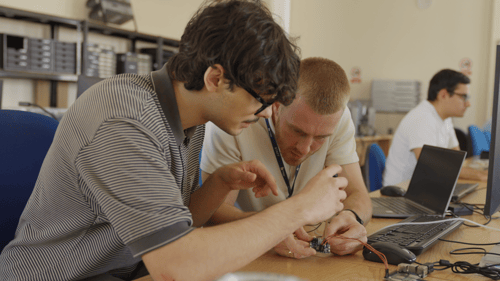A medium shot of two individuals sat at a desk looking intensely at a small PCB with cables coming out of it.